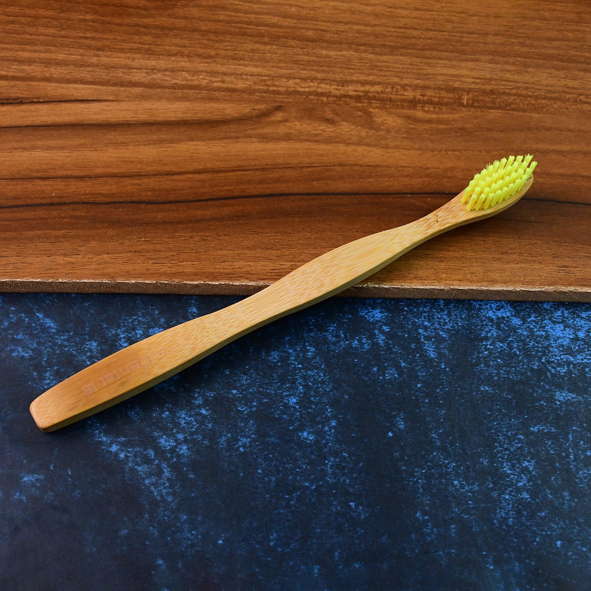 A bamboo toothbrush with yellow bristles is angled on a dark wooden and blue textured background.