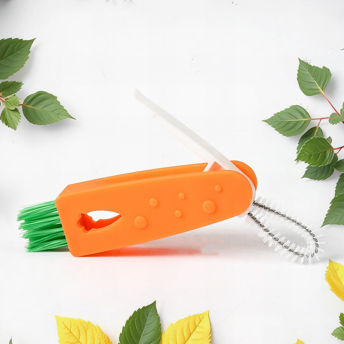 The orange 3-in-1 cleaning brush with green bristles and a white crevice tool is displayed on a white background with leaves.