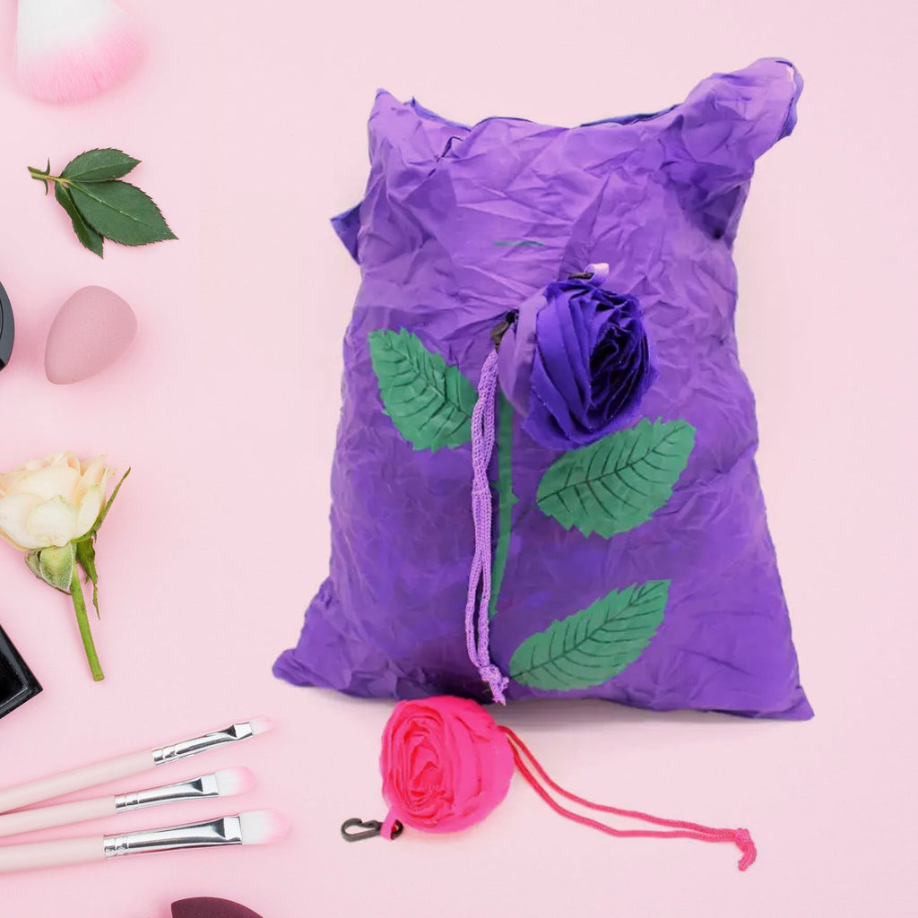 A purple foldable shopping bag with a rose design lies flat next to a pink folded bag on a pink surface.