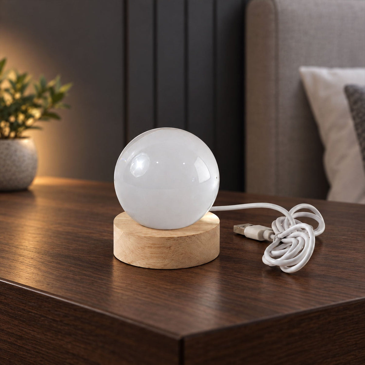 A white spherical lamp on a light wooden base with a coiled white USB cable on a dark wooden table.