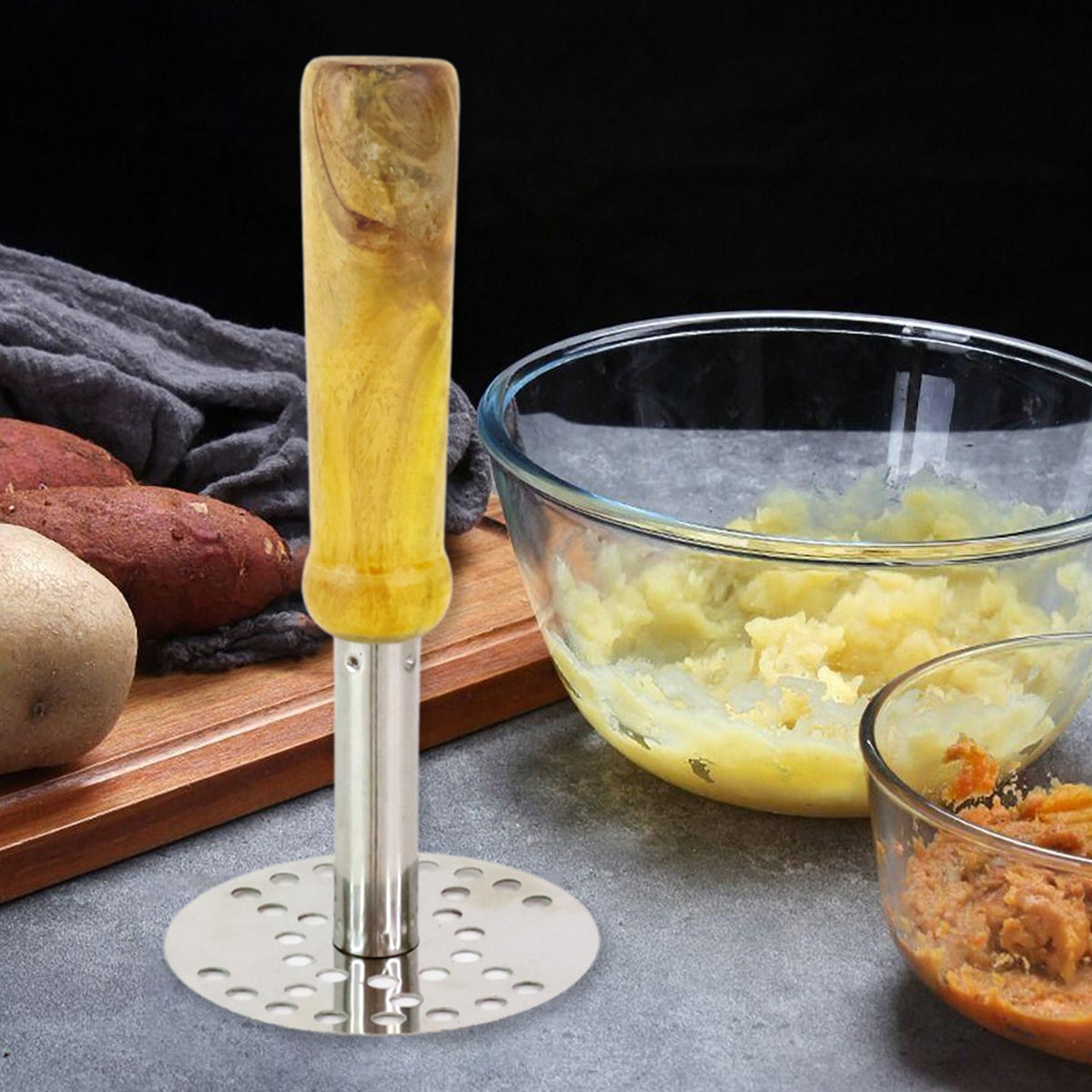 A stainless steel masher with a light wooden handle stands beside bowls of mashed potatoes and sweet potatoes.