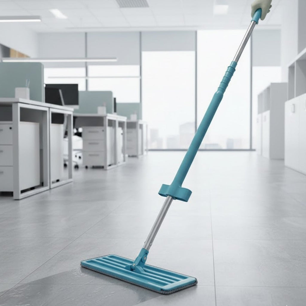 A blue and silver flat mop cleans a grey office floor, with desks and windows in the background.