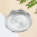 A silver plastic scalloped serving bowl with a textured bottom sits on a two-toned beige and white surface with green leaves