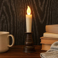 A cream LED candle with a flickering flame sits in a dark bronze holder next to a mug and books.
