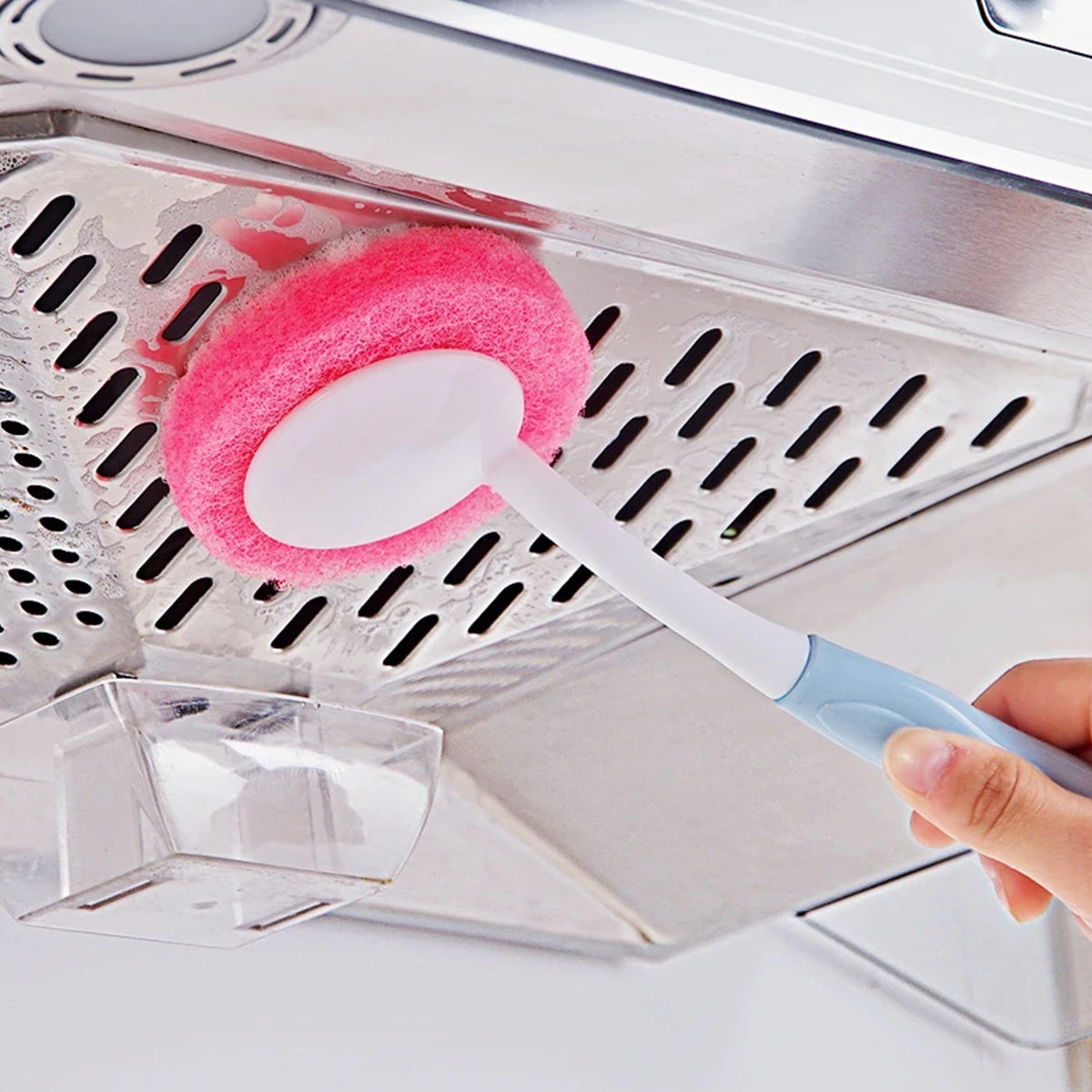 A hand holds a pink and blue long-handled scrubber, cleaning a metallic kitchen hood vent with a pink pad.
