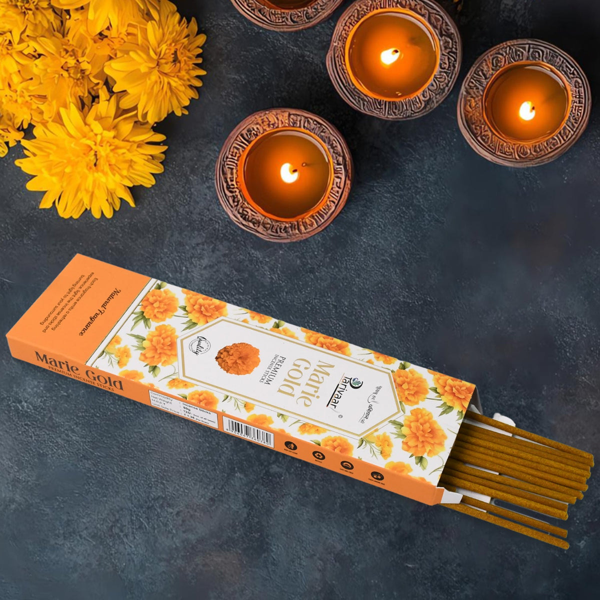 An open orange box of Marie Gold incense sticks with yellow marigolds and three lit diyas.