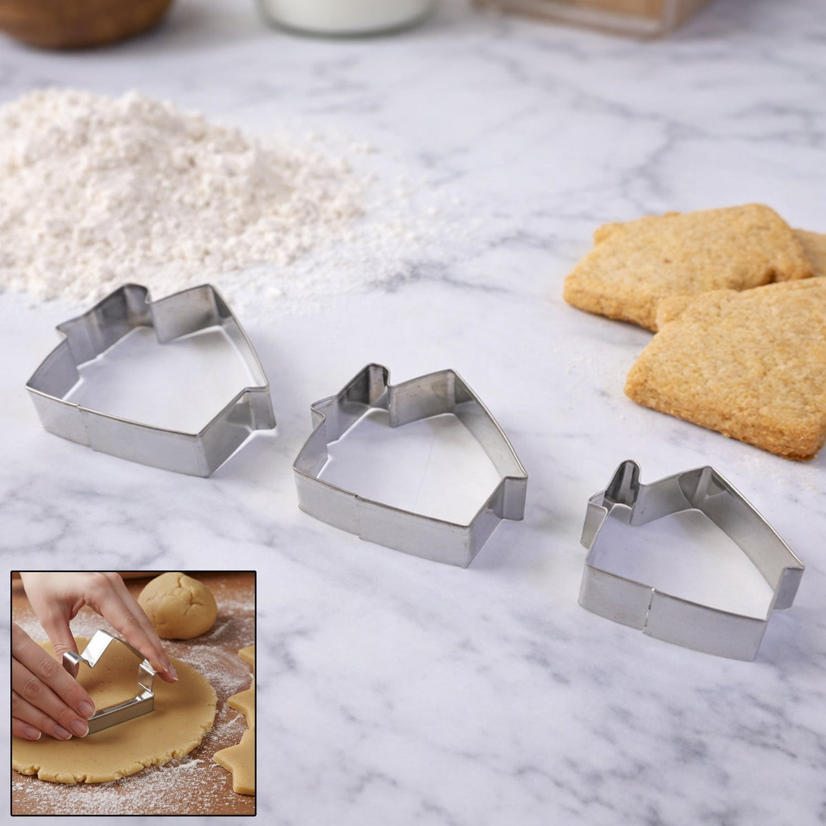 Three house-shaped stainless steel cookie cutters on a marble surface, with flour, baked cookies, and a small inset of a