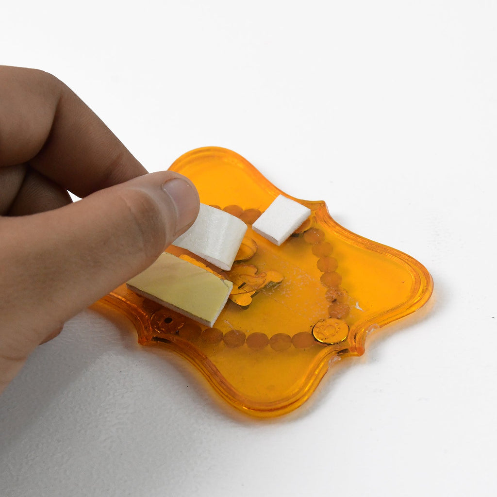 A hand applying white adhesive foam squares to the back of a yellow Shubh Labh wall hanging.