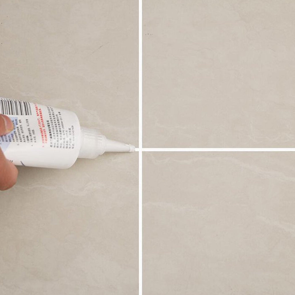 Close-up of a hand applying white sealant from a tube to the intersection of four beige floor tiles.