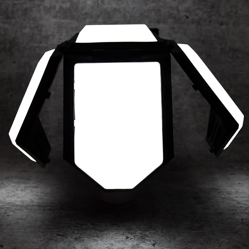The white and black foldable LED lantern with its panels partially open, glowing brightly in a dark setting.