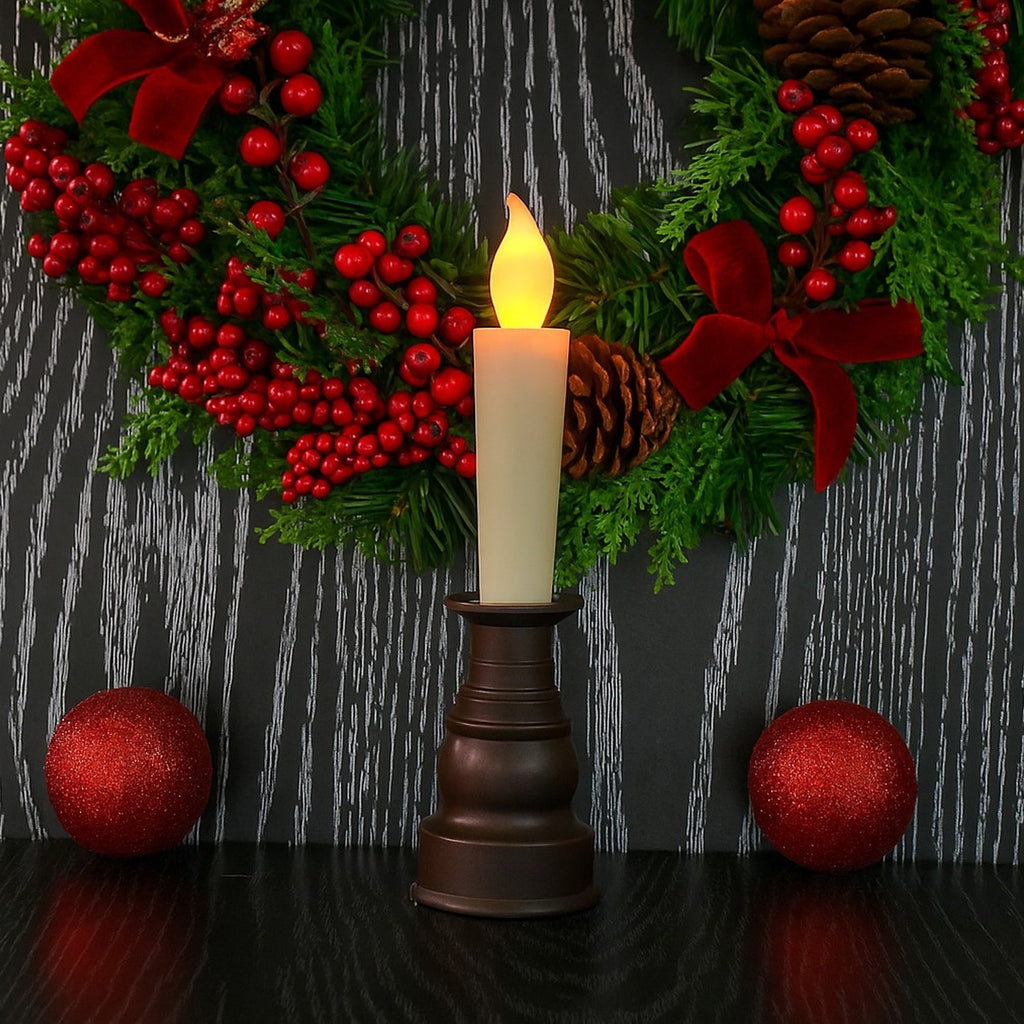 A cream LED candle in a dark bronze holder in front of a festive wreath with red berries and bows.