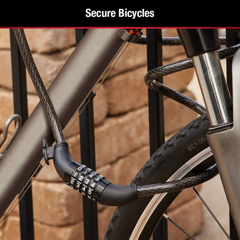 A black cable lock secures a grey bicycle to a metal fence, with a brick wall in the background.