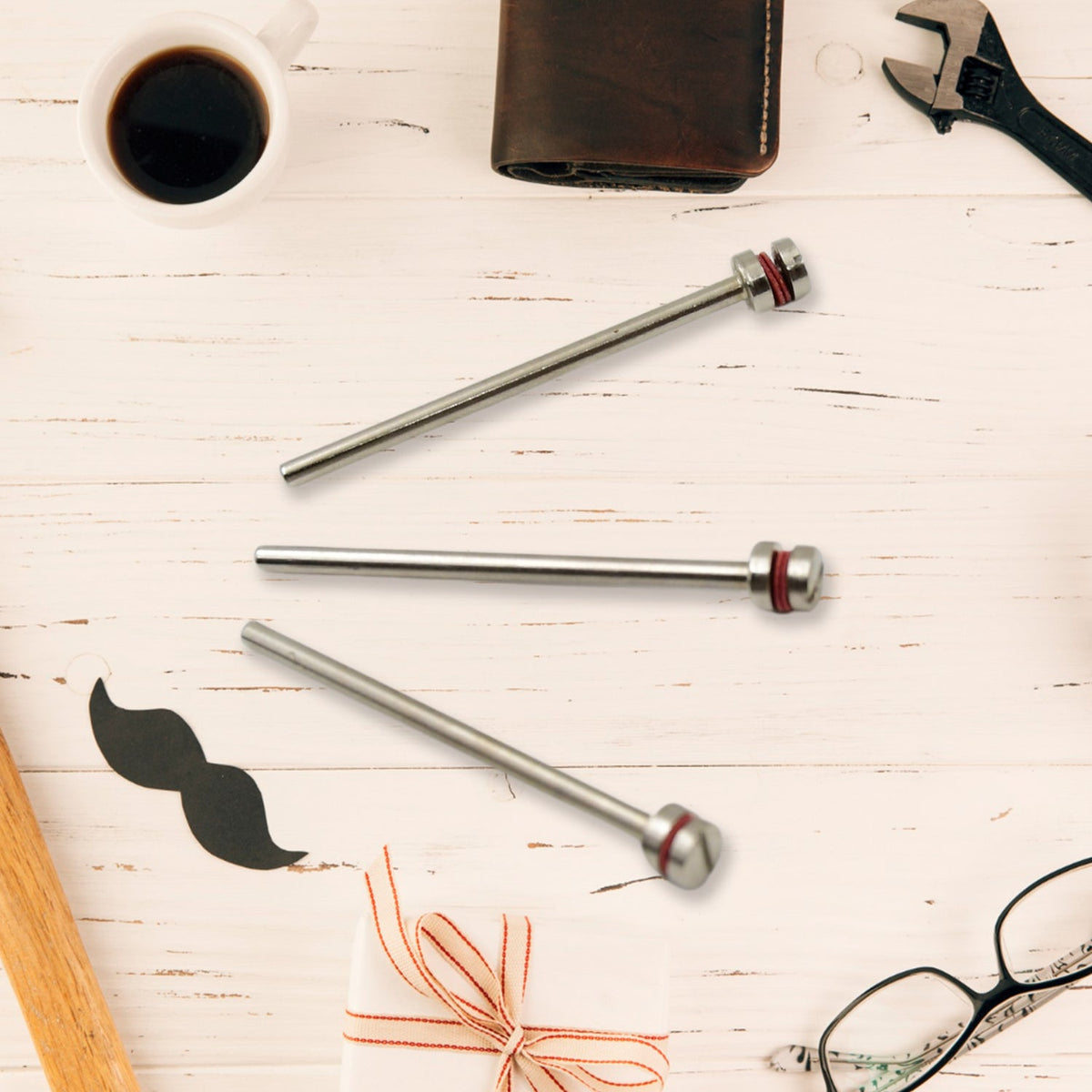Three silver mandrels with red bands are arranged on a light wooden surface with coffee, a wallet, wrench, mustache, and