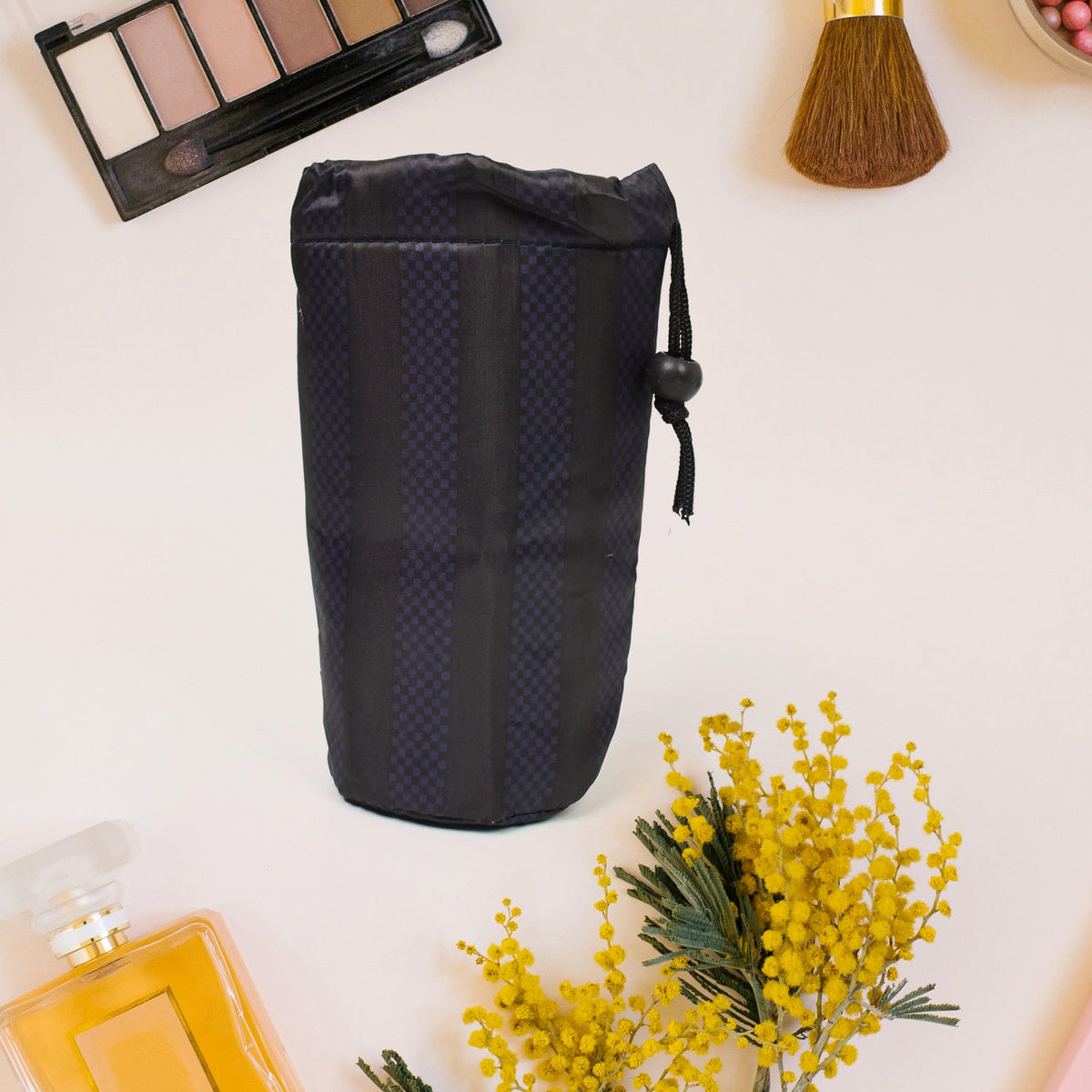 A navy blue drawstring pouch with vertical stripes stands upright on a light surface, surrounded by makeup and a yellow