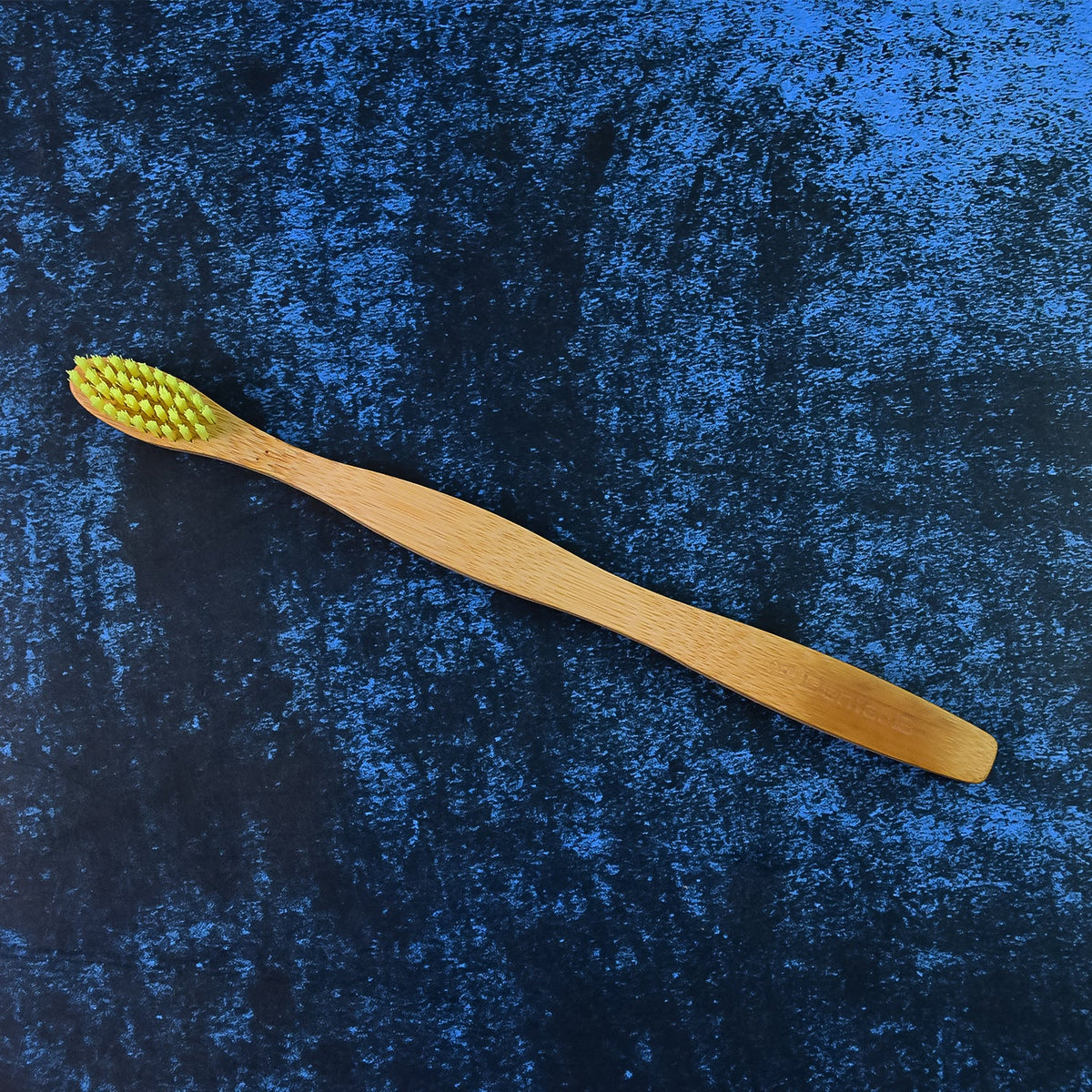 A bamboo toothbrush with yellow bristles is placed diagonally on a dark blue textured surface.