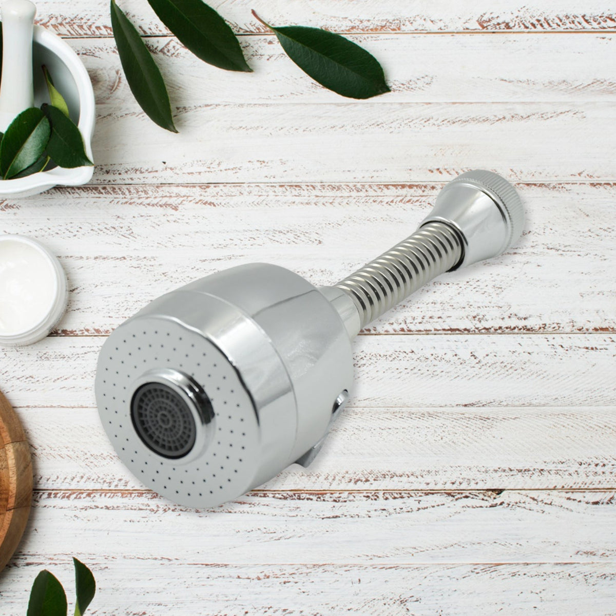 A chrome flexible faucet extension with a sprayer head is on a white wooden surface with green leaves and a mortar and