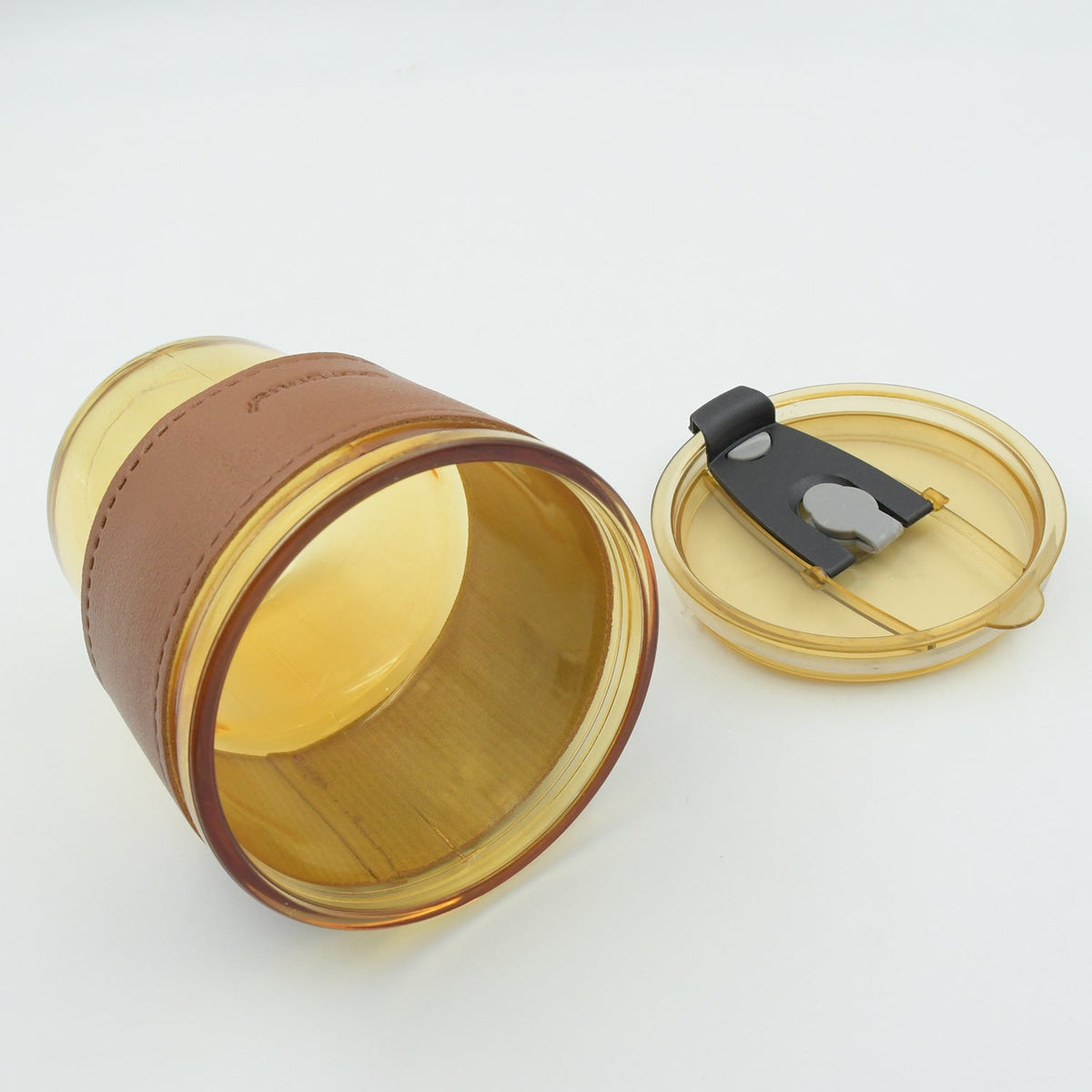 An amber glass cup with a brown leather sleeve lies on its side next to its matching amber lid on a white surface.