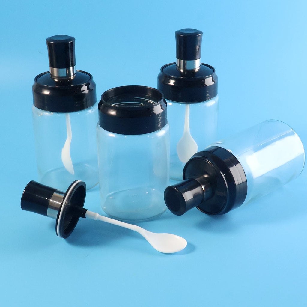 Four transparent spice jars, one with its black lid and white spoon detached on a blue background.