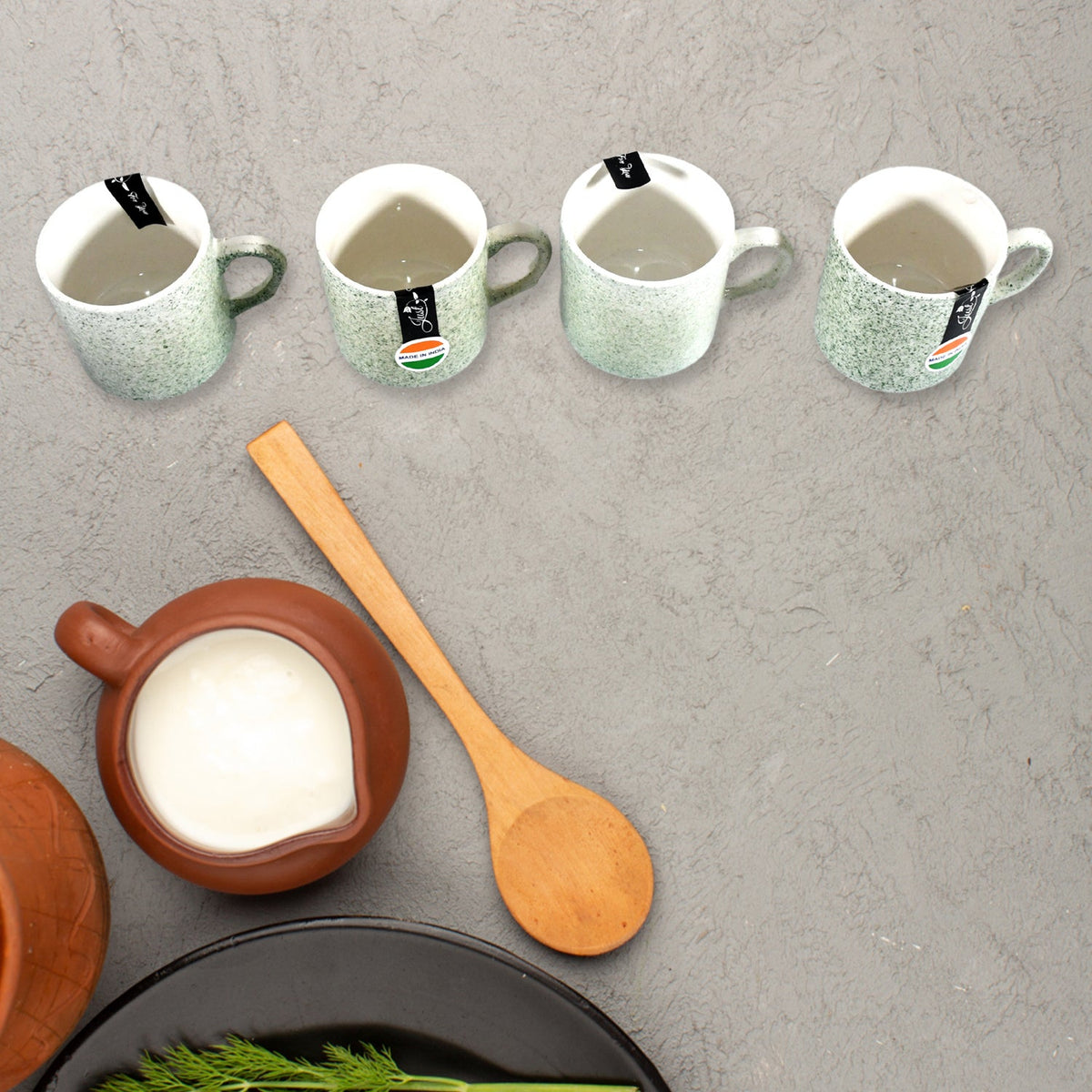 Four speckled gray-green ceramic mugs with handles on a gray background, next to a brown pitcher.