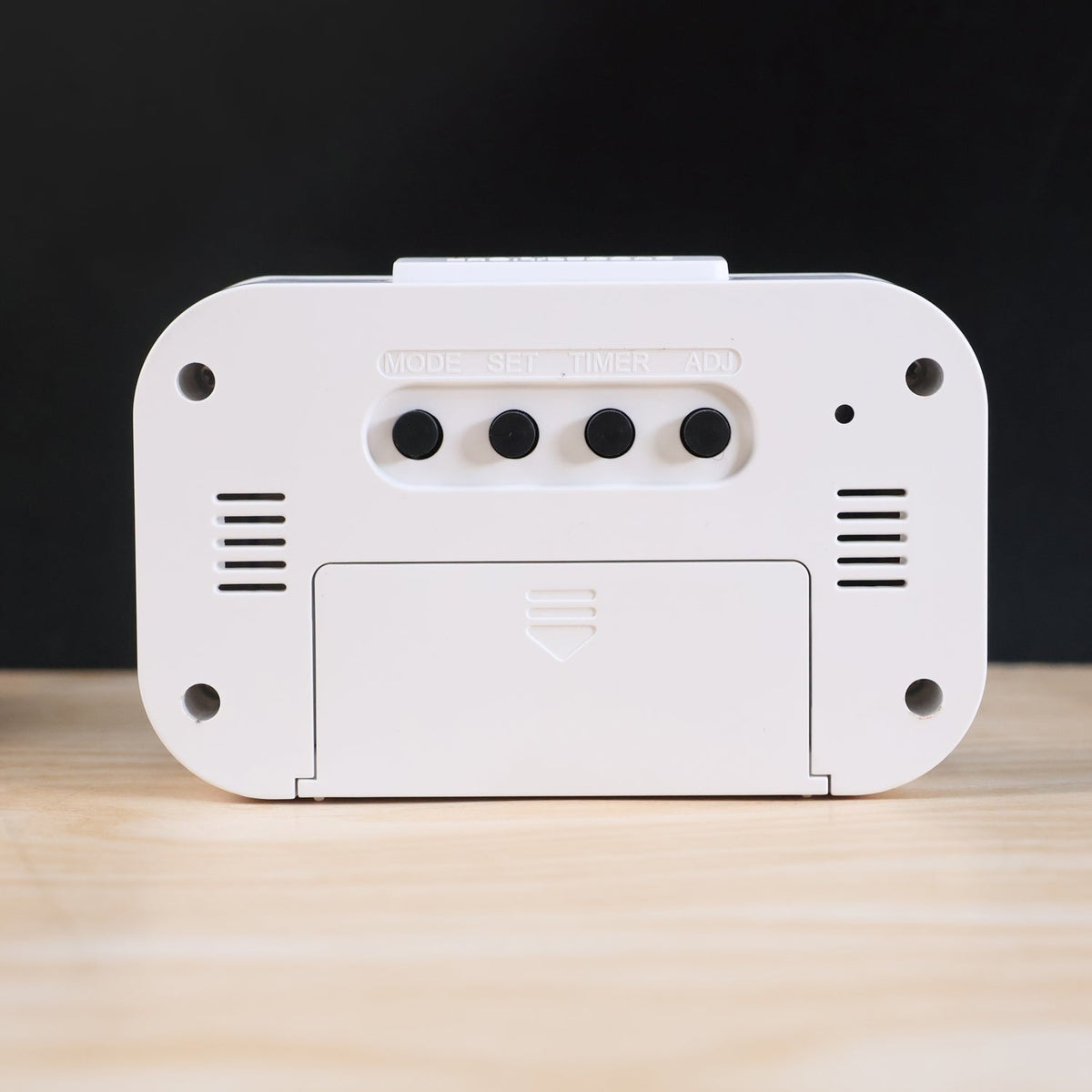 The back of a white digital clock reveals a battery cover and four black control buttons labeled MODE, SET, TIMER, ADJ.