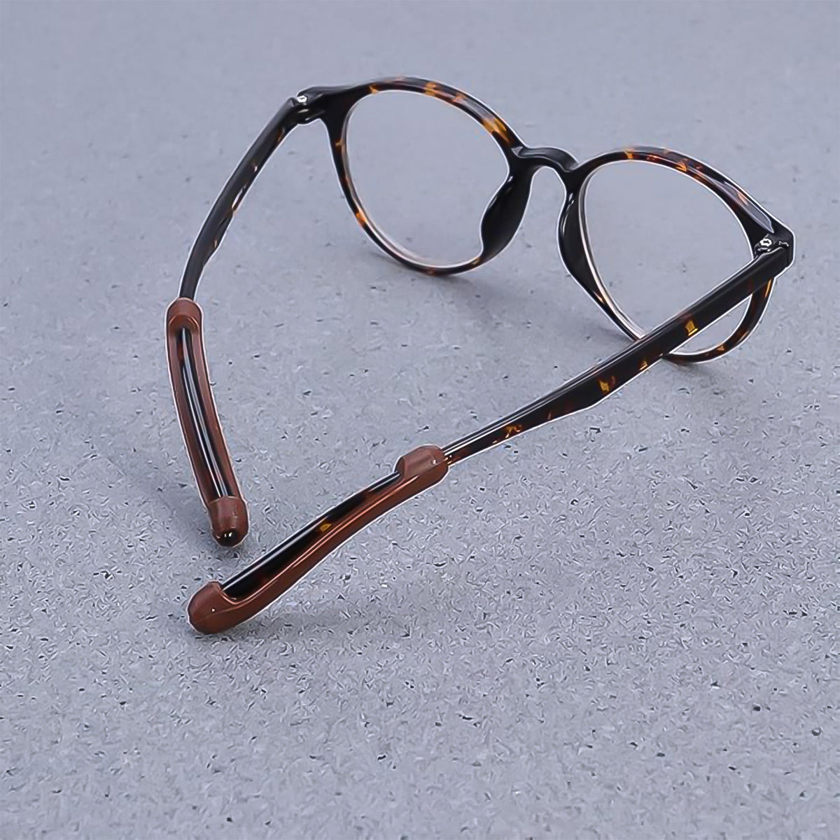Tortoiseshell eyeglasses with brown anti-slip ear grips on the temples, resting on a gray textured surface.