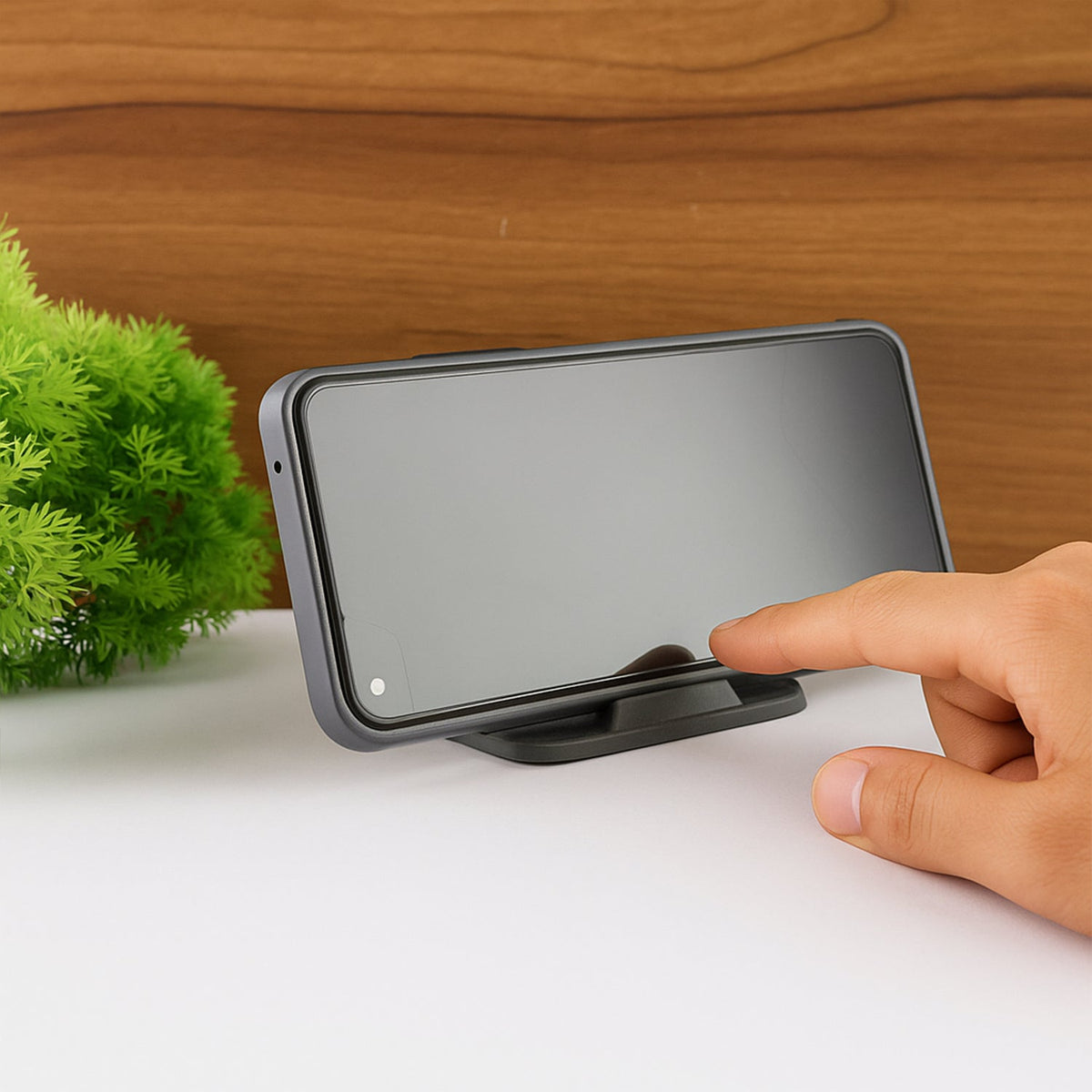 A hand touches the screen of a smartphone resting horizontally on a black mobile stand, next to a green plant.