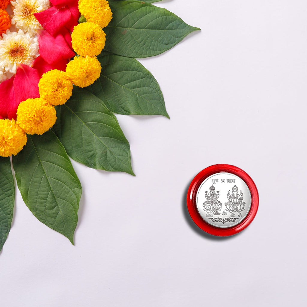 A silver coin with Lakshmi and Ganesha in a red case, beside a floral rangoli design with green leaves and yellow flowers.