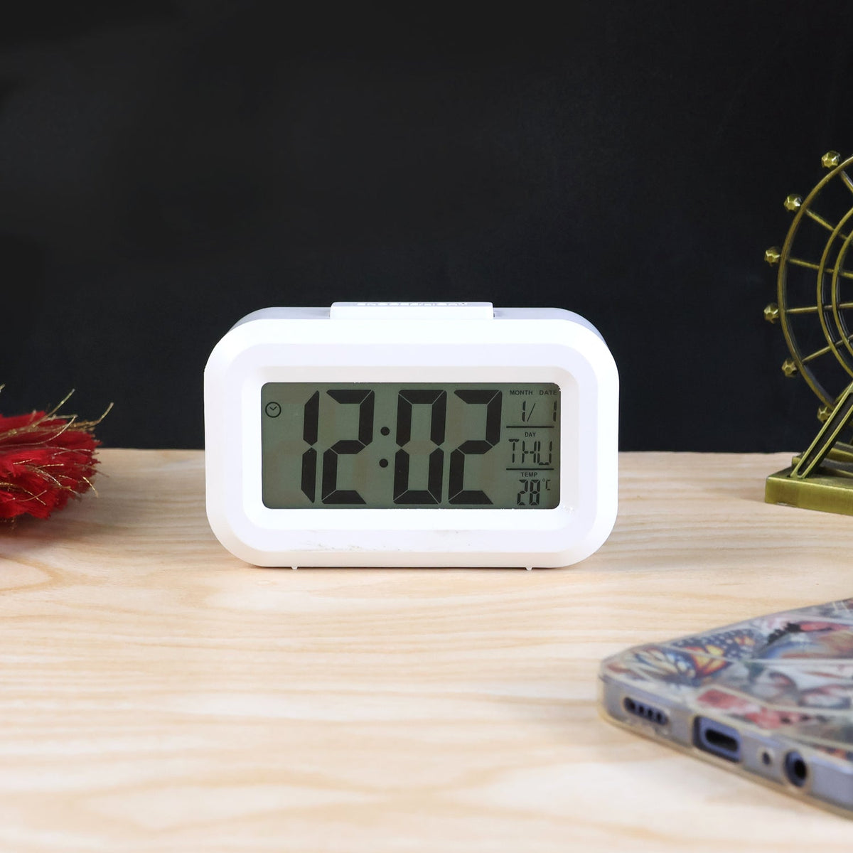 A white digital clock displays 12:02, the date 1/1, THU, and 28 degrees Celsius on a wooden surface.