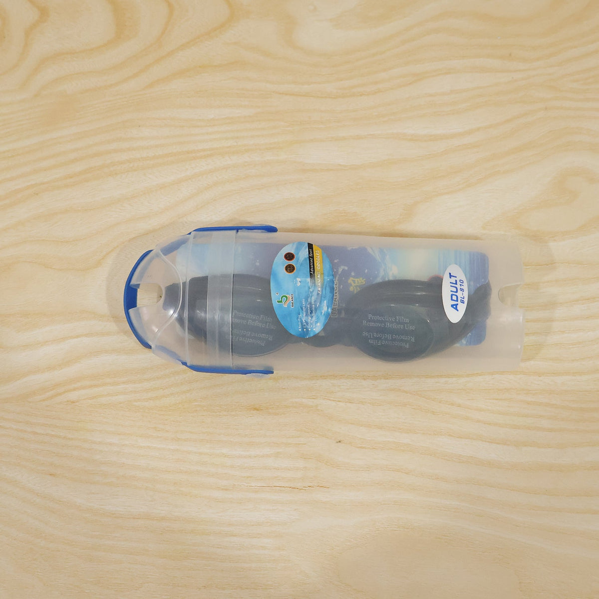 Black swimming goggles are stored in a clear plastic case with a blue lid on a light wooden surface.