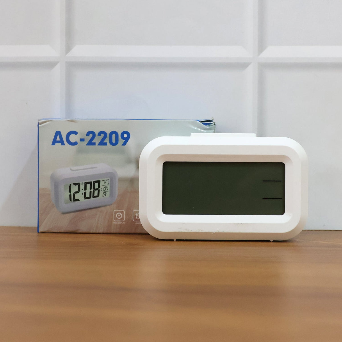 A white digital clock with a dark, blank screen sits next to its blue and white product box.