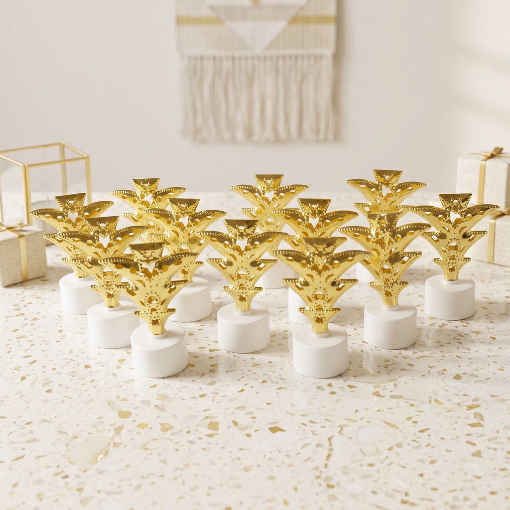A dozen golden Christmas tree LED tealights stand on a white speckled surface, surrounded by gift boxes.