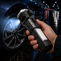 Hand holding a black and silver multi-tool flashlight, illuminating a car tire on a wet street at night.
