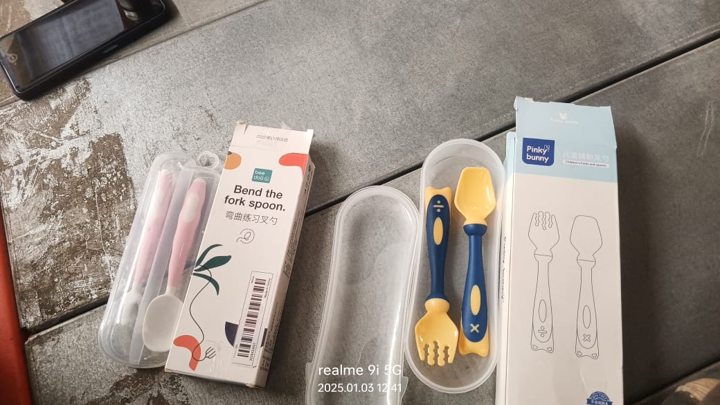 Two sets of baby utensils, one pink and one yellow/blue, in packaging on a dark surface next to a phone.