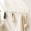 Six transparent adhesive hooks with gold borders holding keys, a towel, and a brush on a white wall.