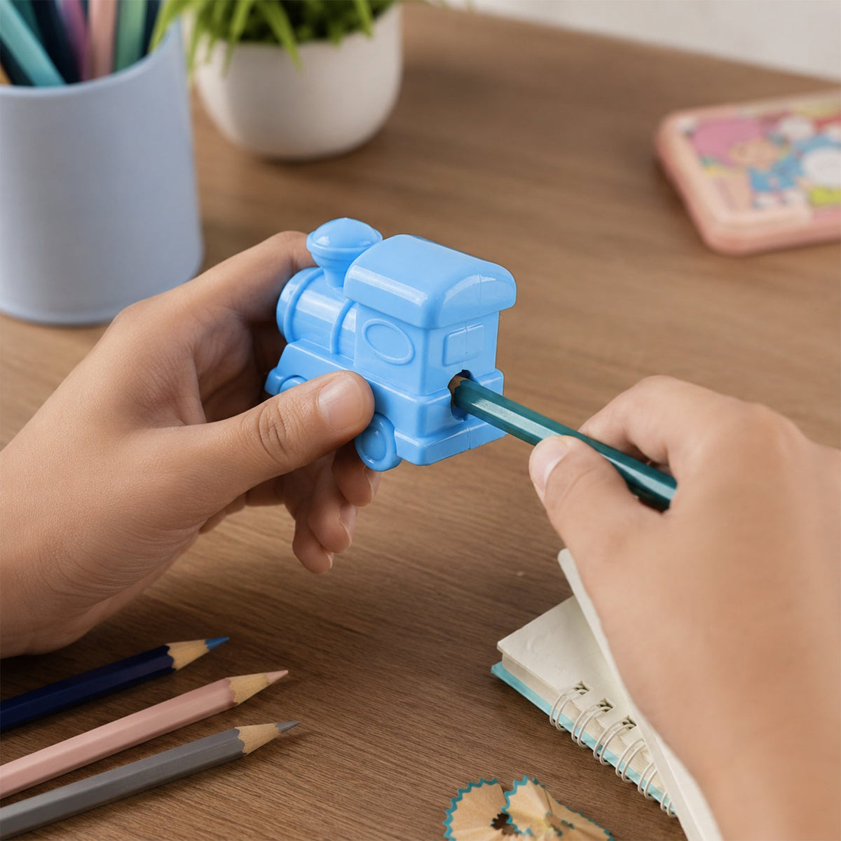 Hands sharpening a green pencil with a light blue train-shaped sharpener on a wooden desk with colored pencils.
