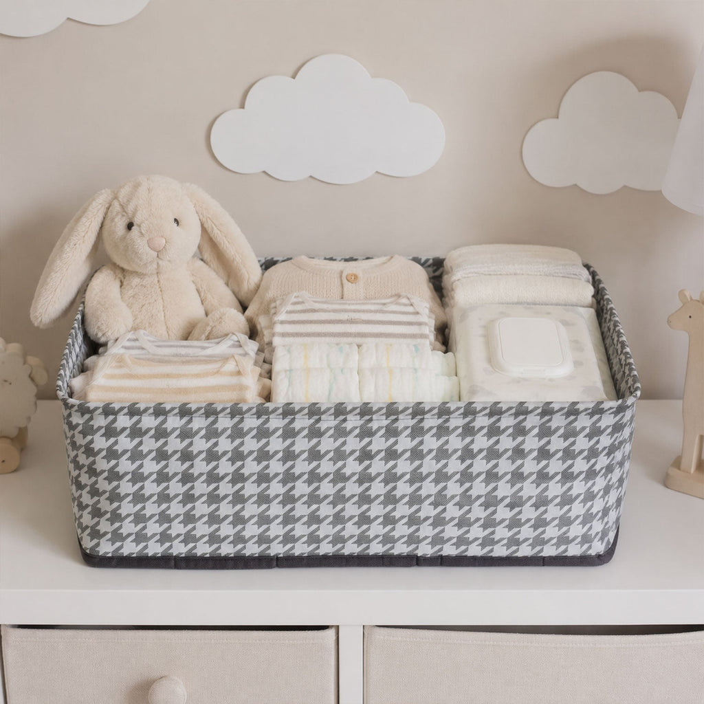 A gray and white houndstooth fabric basket holds baby clothes, diapers, wipes, and a plush bunny on a white dresser.