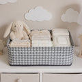 A gray and white houndstooth fabric basket holds baby clothes, diapers, wipes, and a plush bunny on a white dresser.
