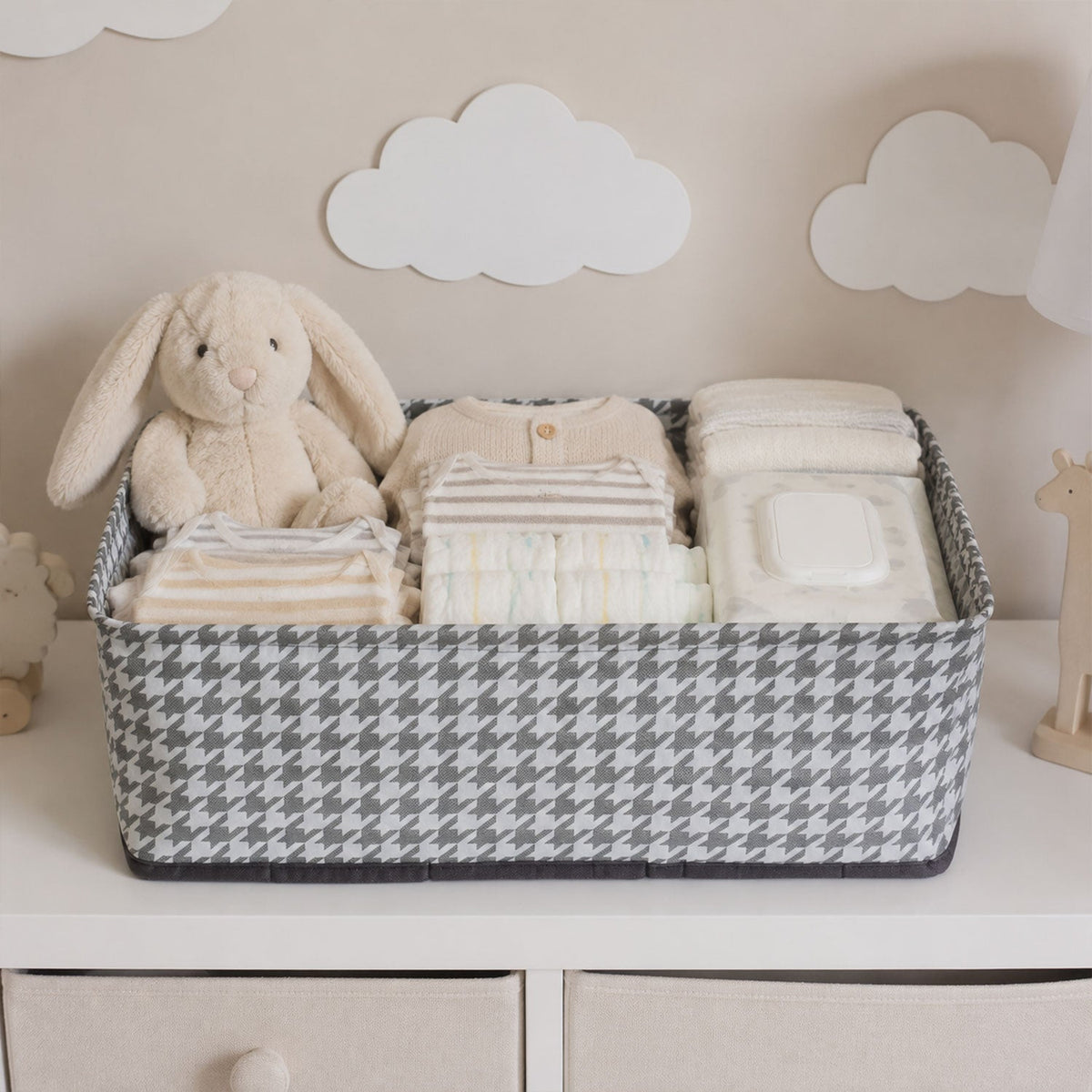 A gray and white houndstooth fabric basket holds baby clothes, diapers, wipes, and a plush bunny on a white dresser.
