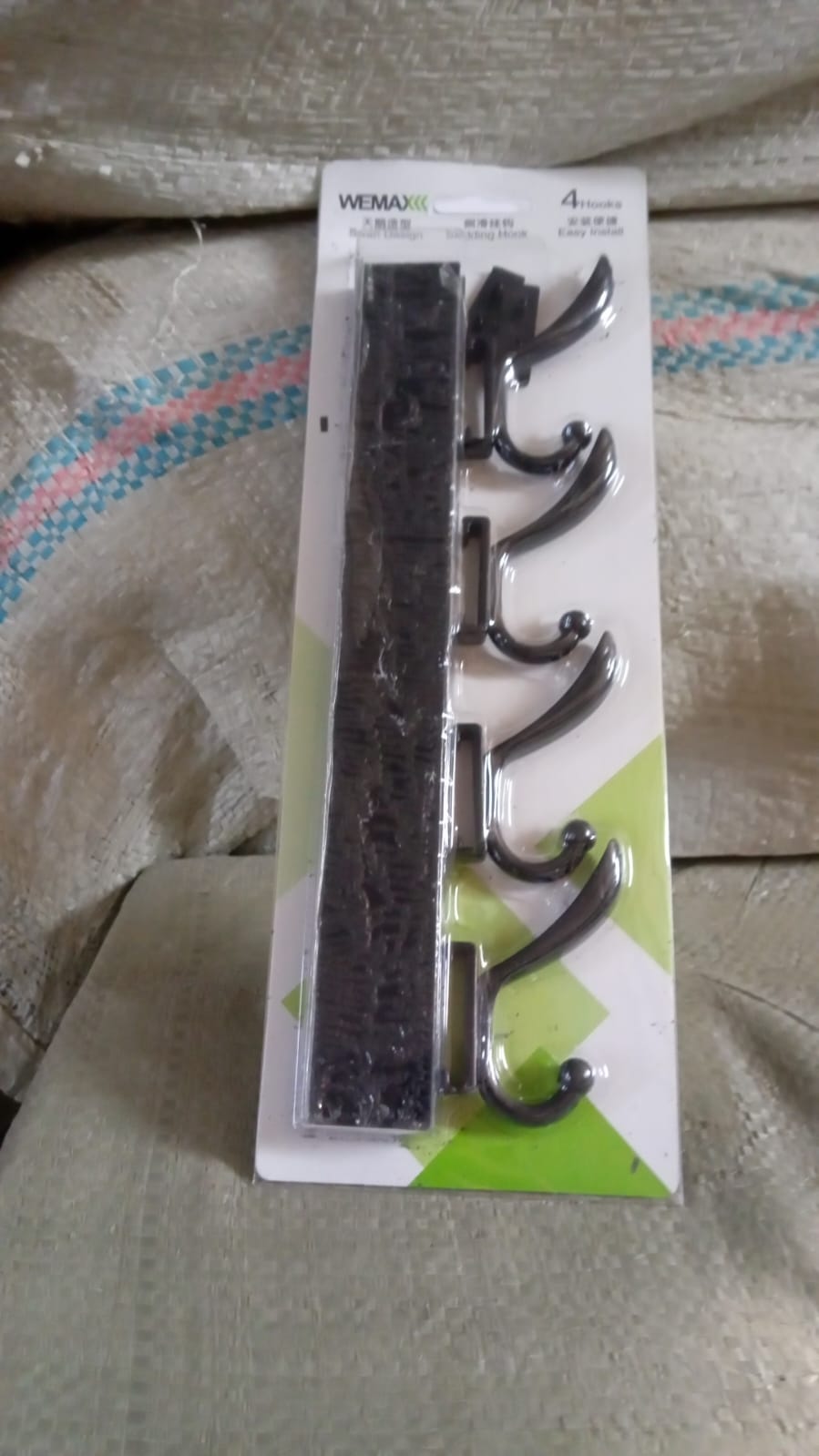 Black four-hook coat rack in its clear plastic packaging with a white and green label, resting on a textured surface.