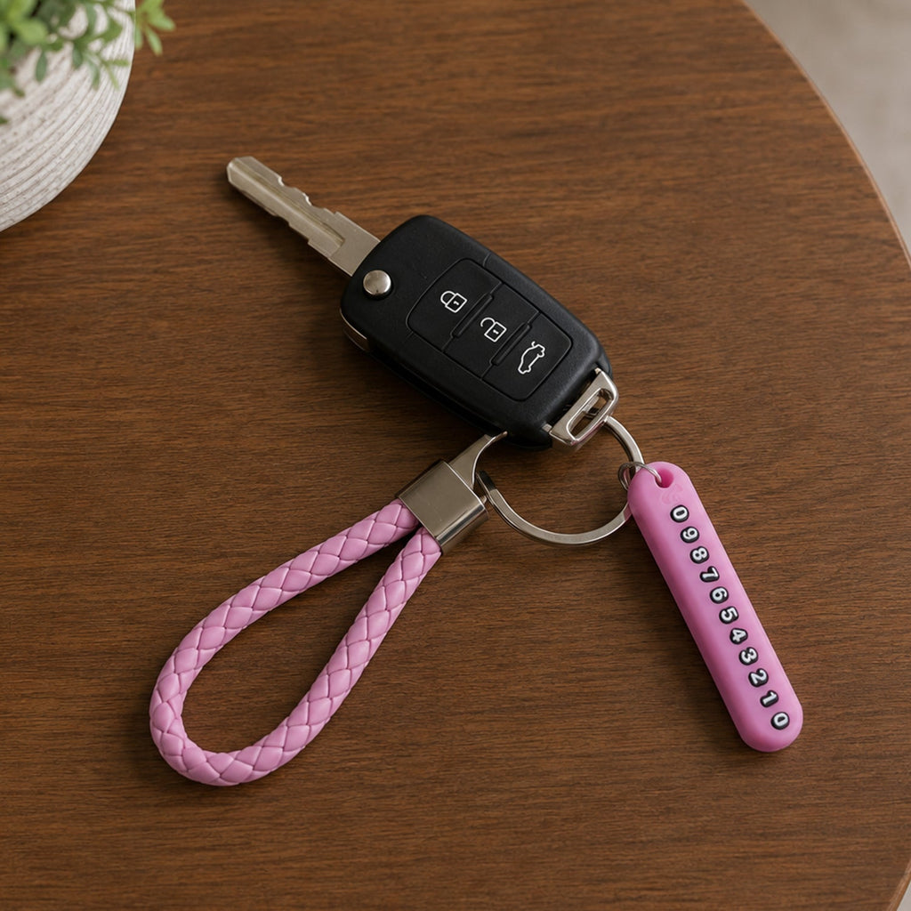 A pink braided wrist loop keychain with a car key and a pink number tag on a wooden table, with a plant in the background.