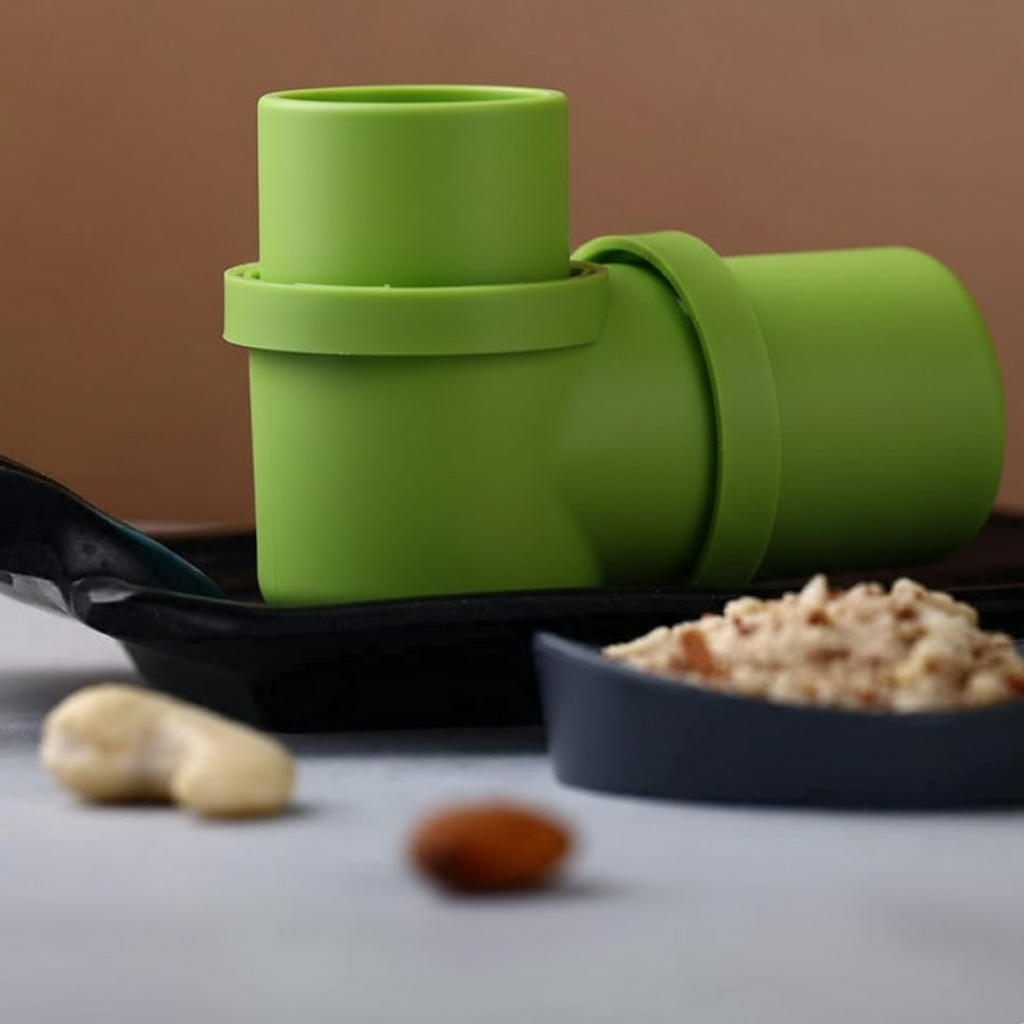 A green dry fruit slicer rests on a dark tray next to a small bowl of sliced nuts and a whole cashew.