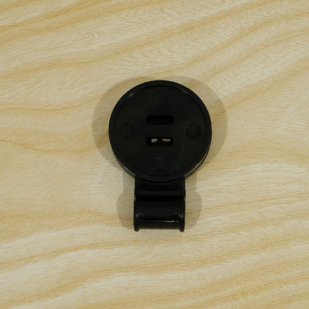 A single black plastic shade cloth clip, closed, lying flat on a light wood grain surface.