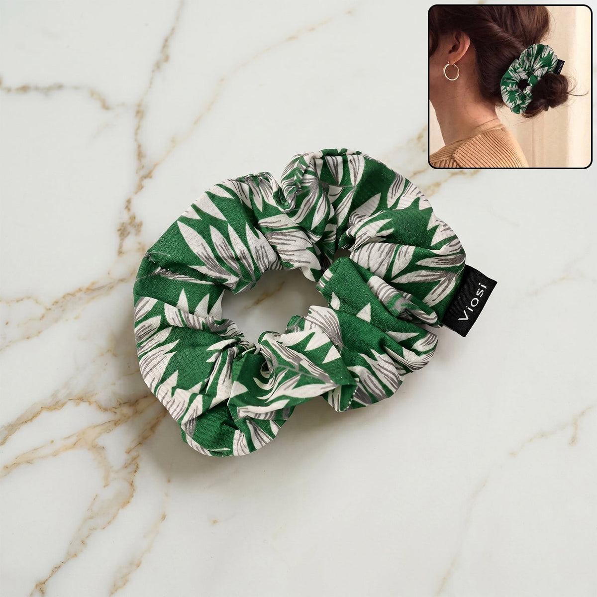 A green and white leaf print scrunchie with a black 'viosi' tag rests on a marble surface, with an inset showing it in a bun.
