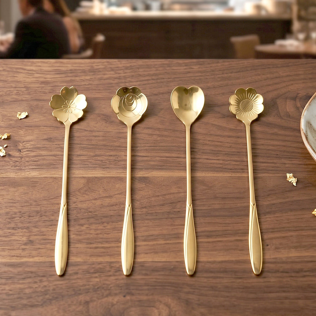 Four gold decorative spoons with flower and heart shapes are displayed on a wooden table with gold flakes.