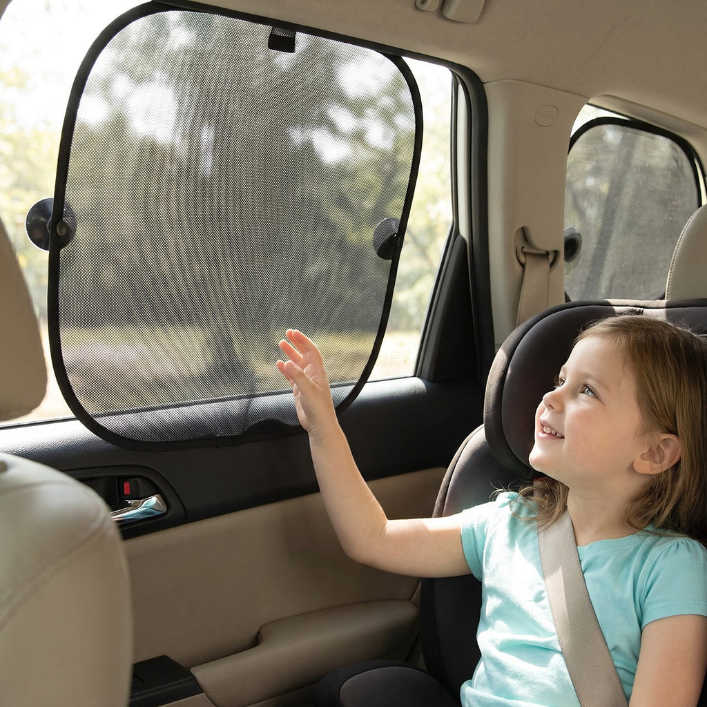 A young girl in a car seat reaches towards a black mesh foldable sunshade with suction cups on the car window.
