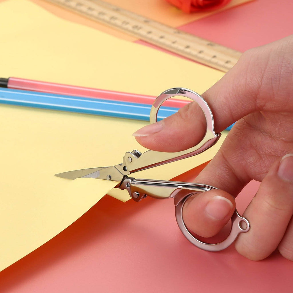 A hand uses open silver folding scissors to cut yellow paper, with a ruler and pencils in the background.