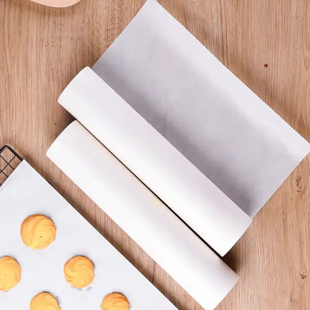 Two rolls of white non-stick baking paper on a wooden surface next to a baking sheet with cookies on a piece of the paper.