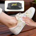 A hand uses a white wet wipe to clean yellow splatters off a beige canvas shoe on a wooden bench.