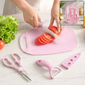 A person slices red tomatoes on a pink cutting board with a matching pink knife, alongside a peeler and scissors.