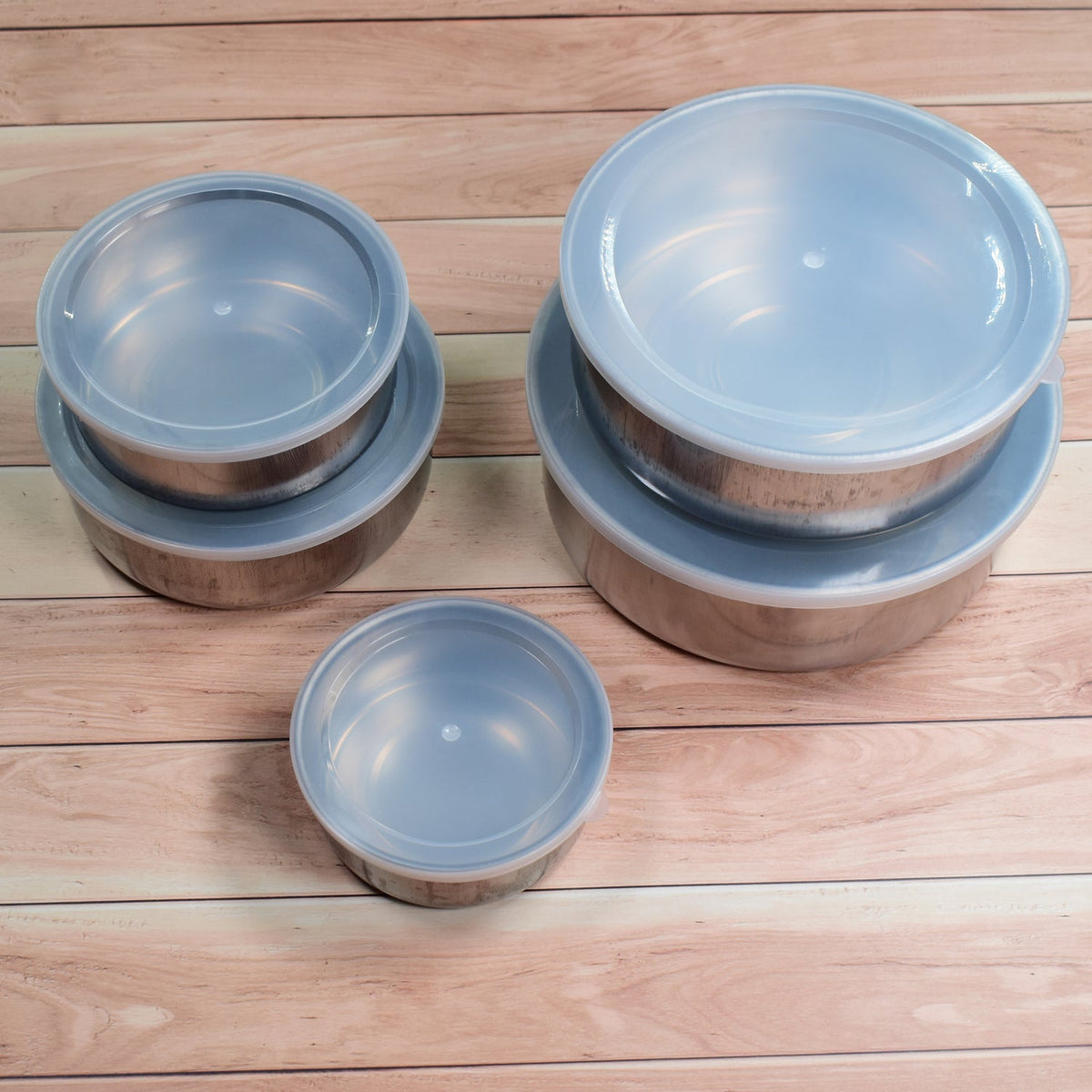 Two stacks of stainless steel food containers with clear lids, and one single container, arranged on a light wooden surface.
